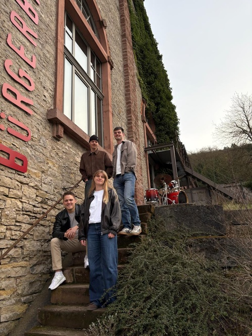 Das Bild zeigt vier Personen, die vor einem alten Gebäude mit der Aufschrift "BURGER" auf der Wand stehen. Drei Personen stehen auf den Stufen, eine sitzt. Im Hintergrund sind Trommeln zu sehen. Die Umgebung wirkt rustikal und natürlich.