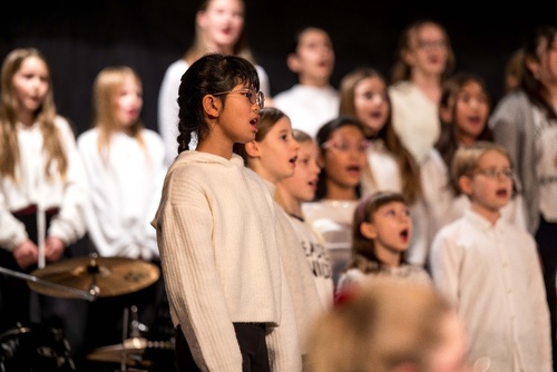 Das Bild zeigt einen Chor von Kindern, die gemeinsam singen. Sie tragen weiße Oberteile und stehen in mehreren Reihen. Eine Person mit Brille und Zopf scheint zu dirigieren oder mitzusingen. Im Hintergrund sind weitere Kinder unscharf zu erkennen.